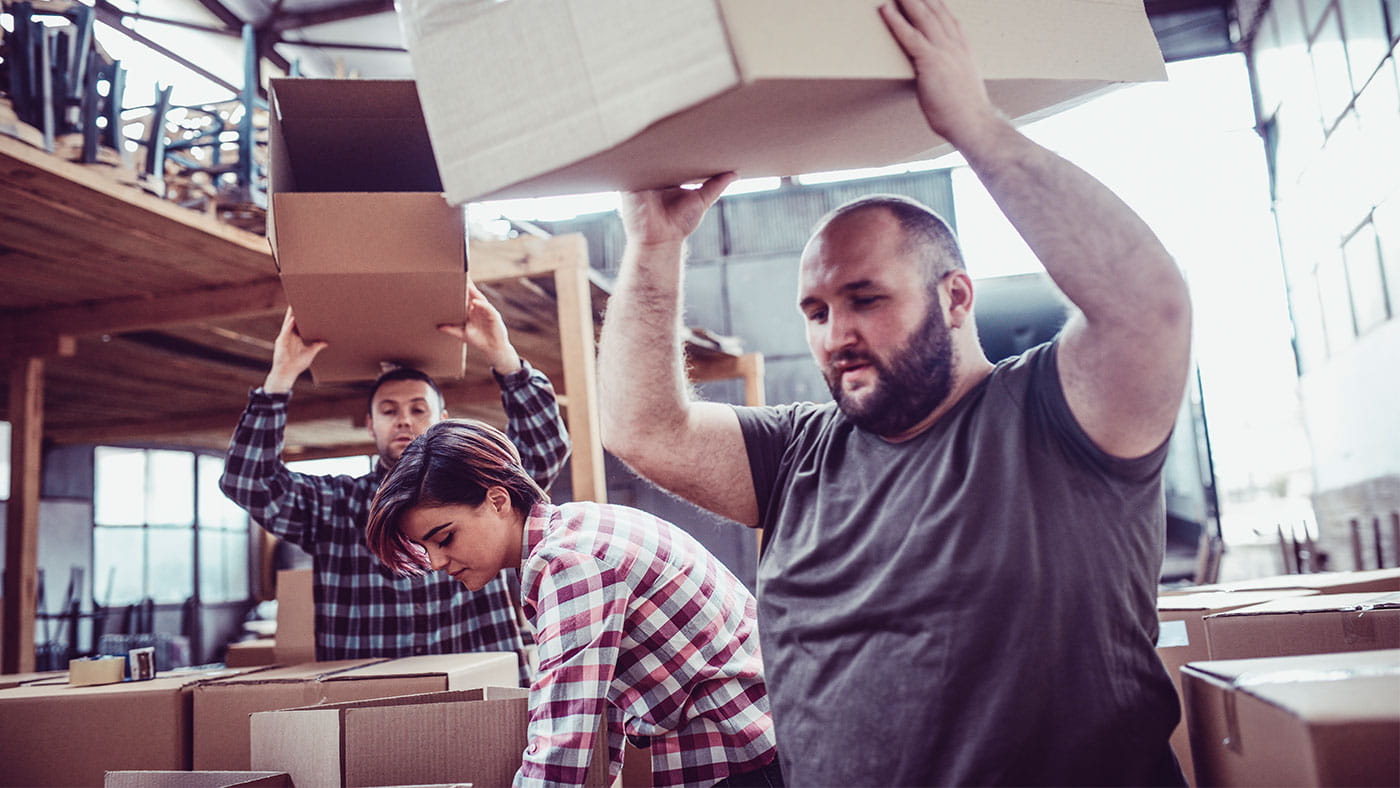 Two men and one woman in a warehouse with the men carrying cardboard boxes over their heads.