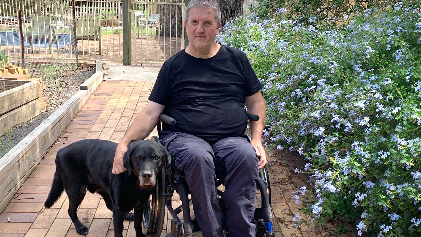 Mick is in a wheelchair outside with his black Labrador Dino.