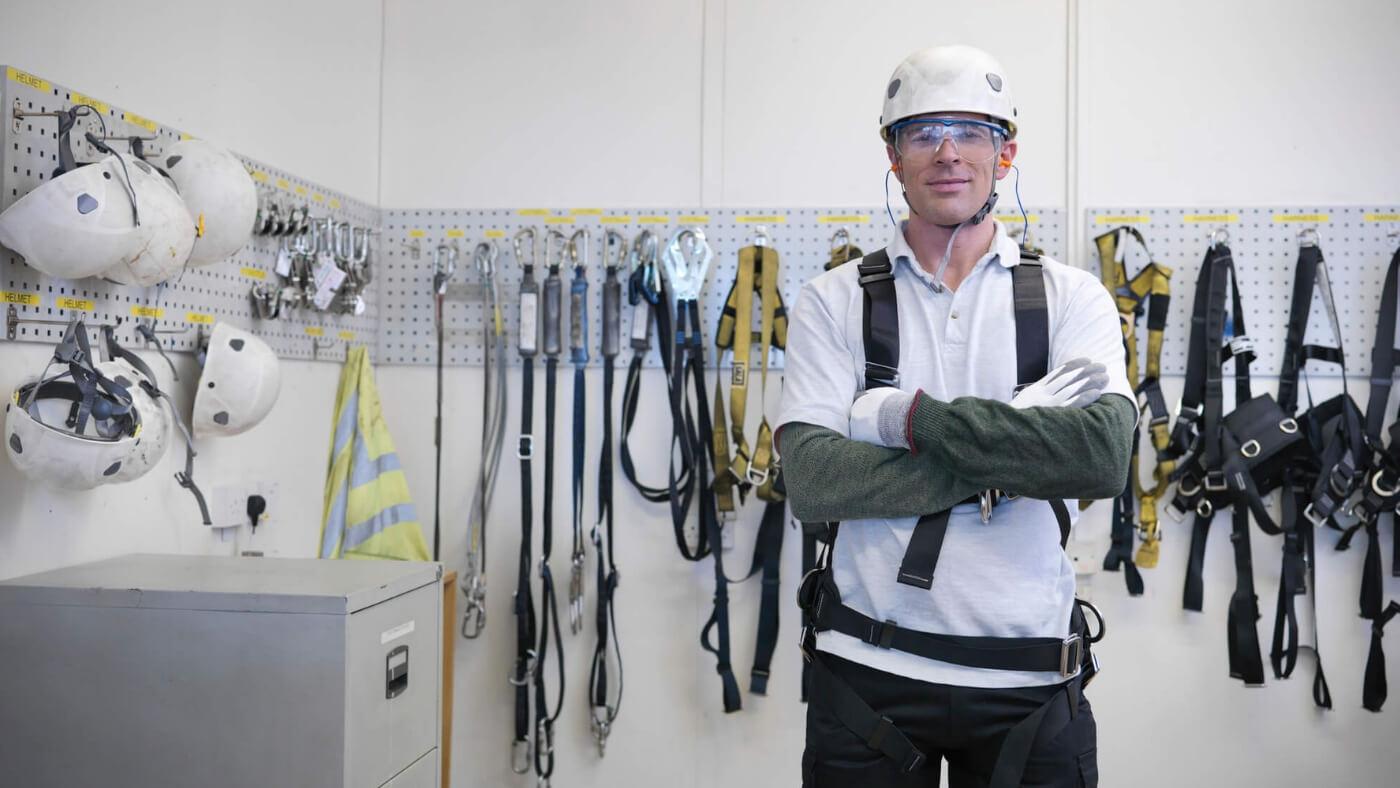 Man standing with arms crossed wearing a helmet, safety glasses, a harness in a room full of ropes and helmets