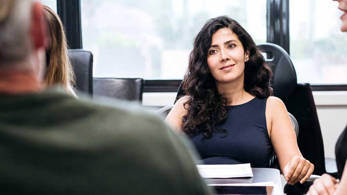 A lady with long dark curly hair in formal wear leans back in her chair at a meeting focused on a colleague who is to the left of the camera.