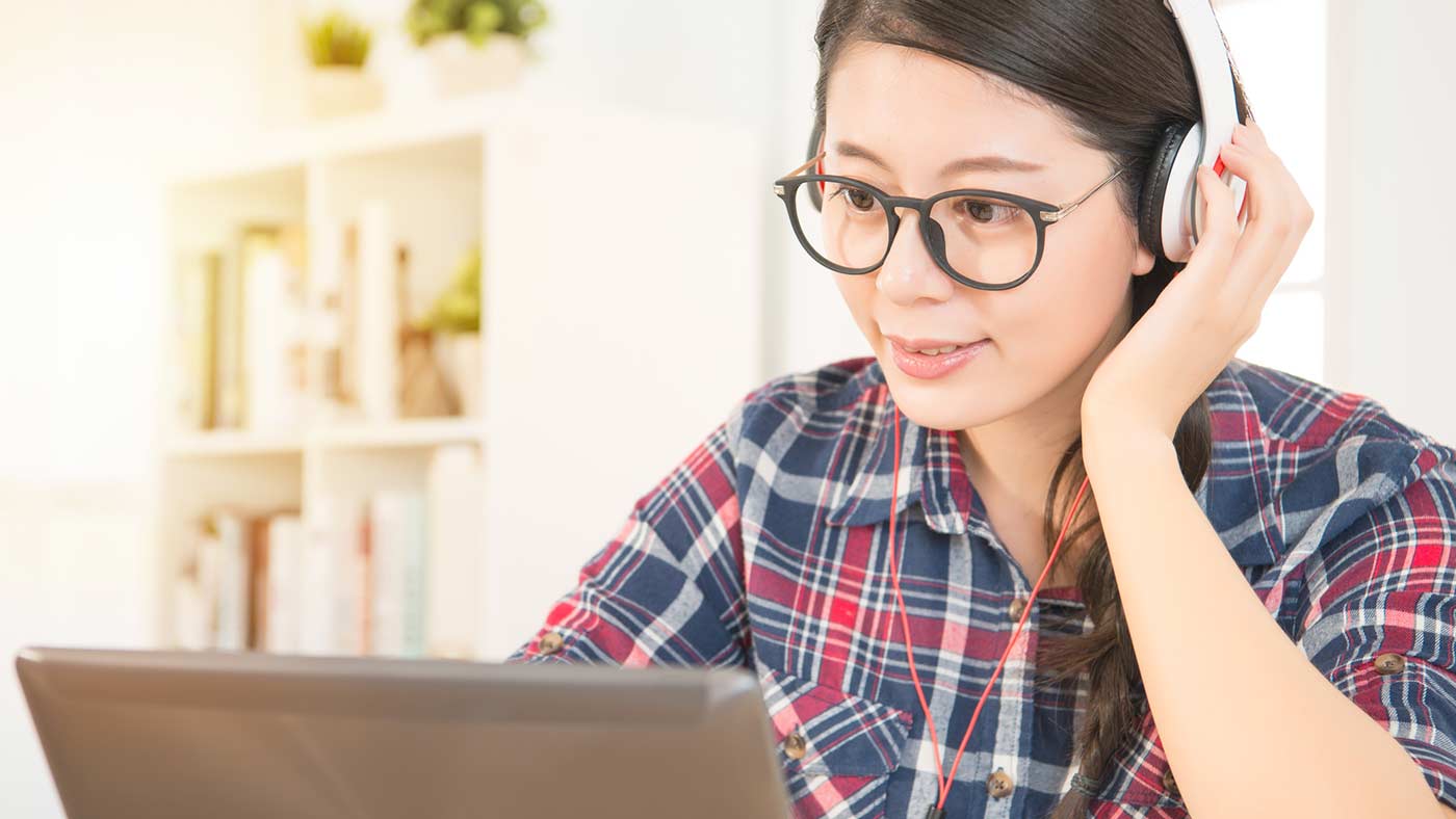 A lady with glasses on is looking at a laptop with headphones on.