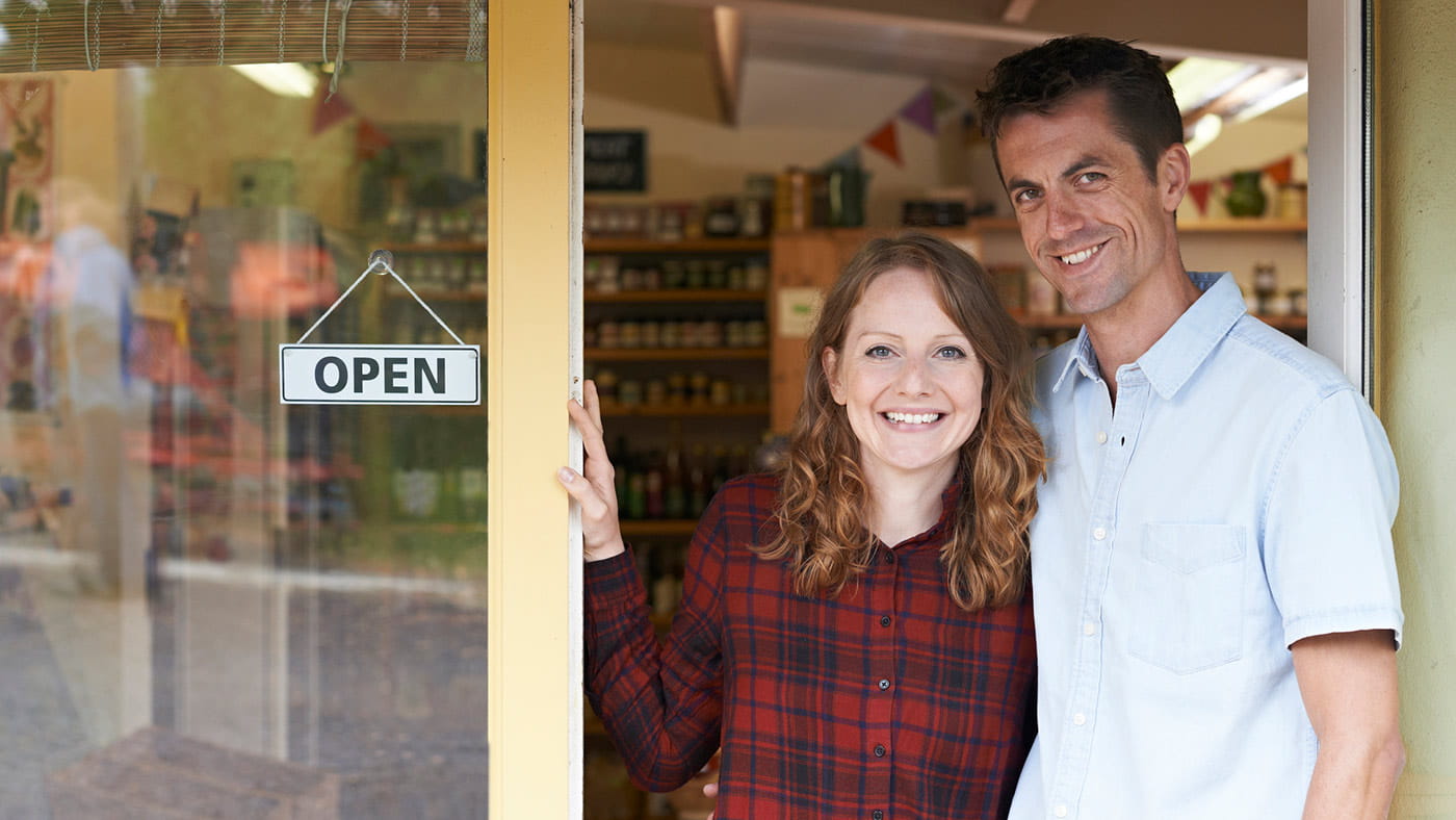 Couple standing at the entrance to their store next to an open for business sign