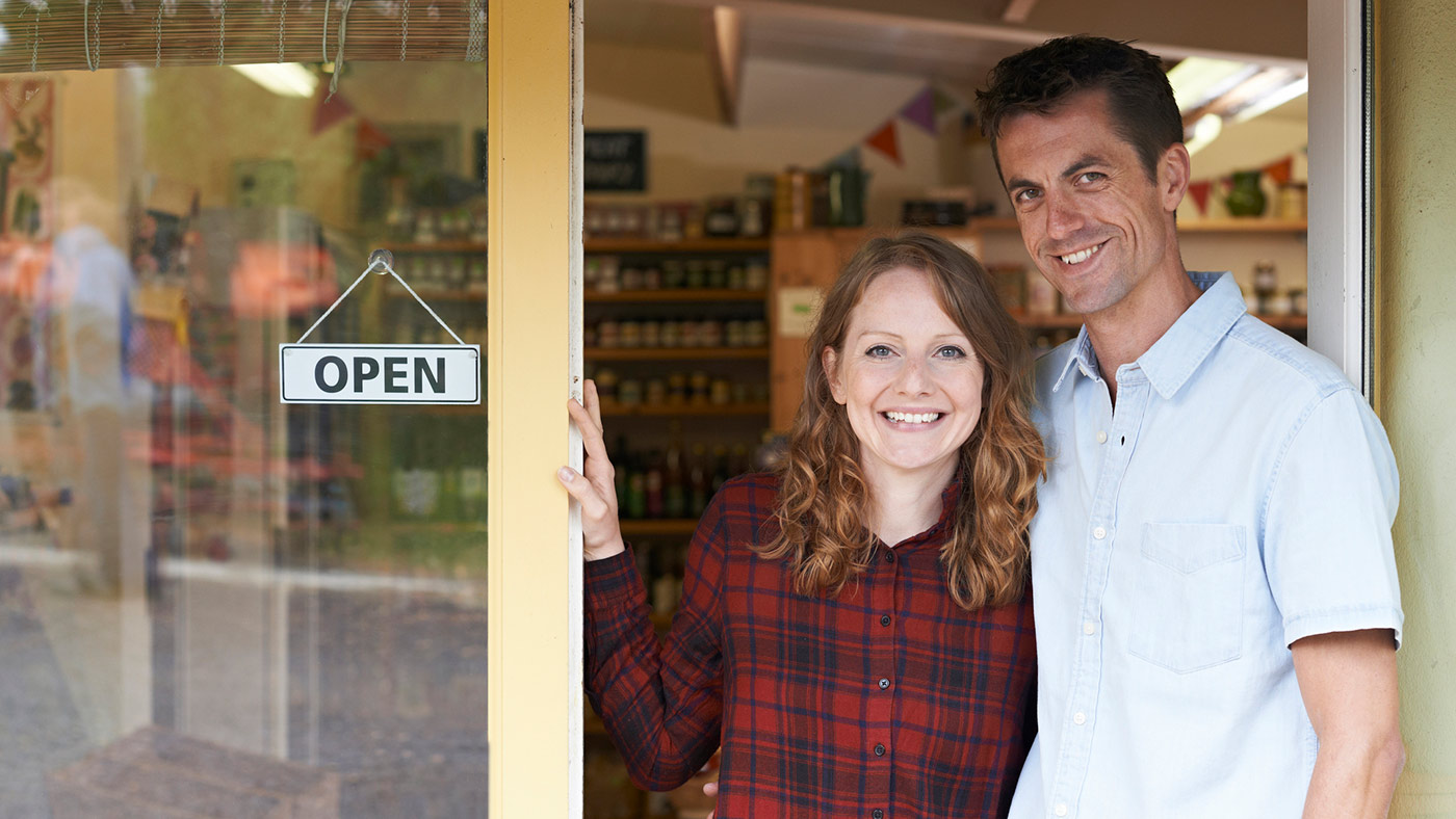 Couple standing at the entrance to their store next to an open for business sign