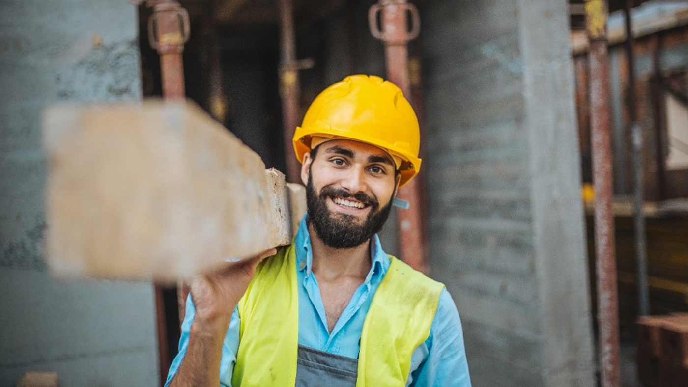 Smiling construction worker wearing a hard hat and high visibility vest carries a plank of wood.