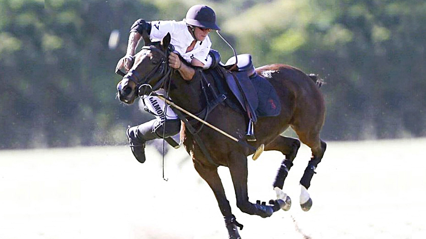Angus Gilmore a polo player is lifting himself onto a horse on a polo field.