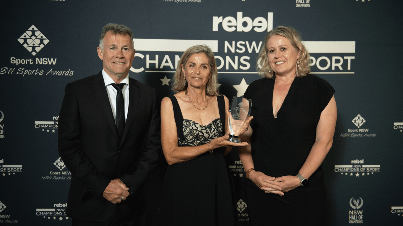Three people standing in front of a media banner with logos of NSW Champions of Sport, NSW Hall of Champions and Sport NSW NSW Sports Awards. The middle person holds a trophy.