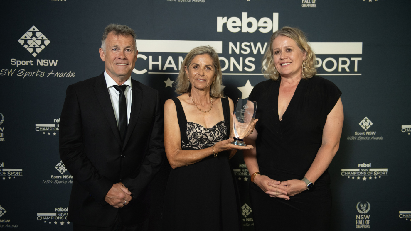 Three people standing in front of a media banner with logos of NSW Champions of Sport, NSW Hall of Champions and Sport NSW NSW Sports Awards. The middle person holds a trophy.