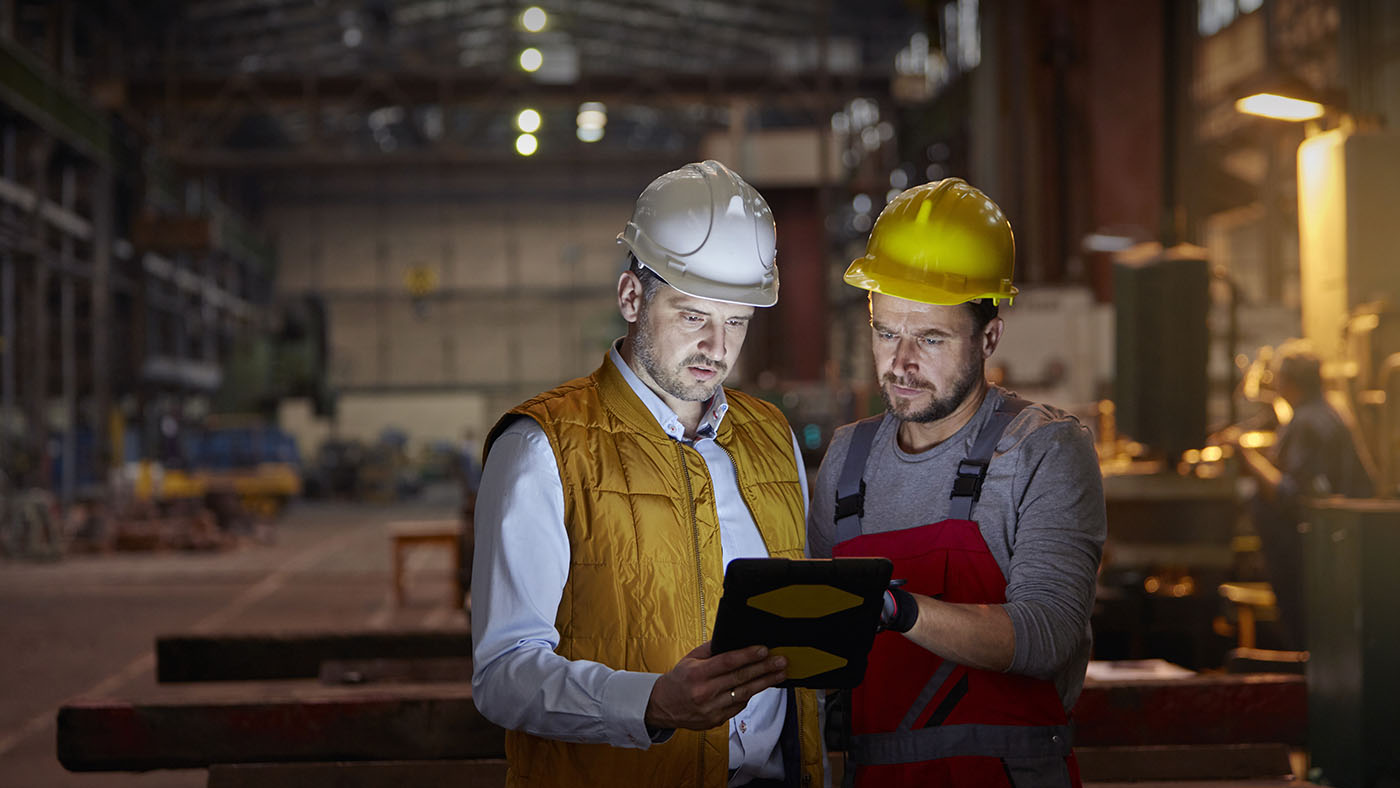Two men in safety gear look at ipad in factory setting.