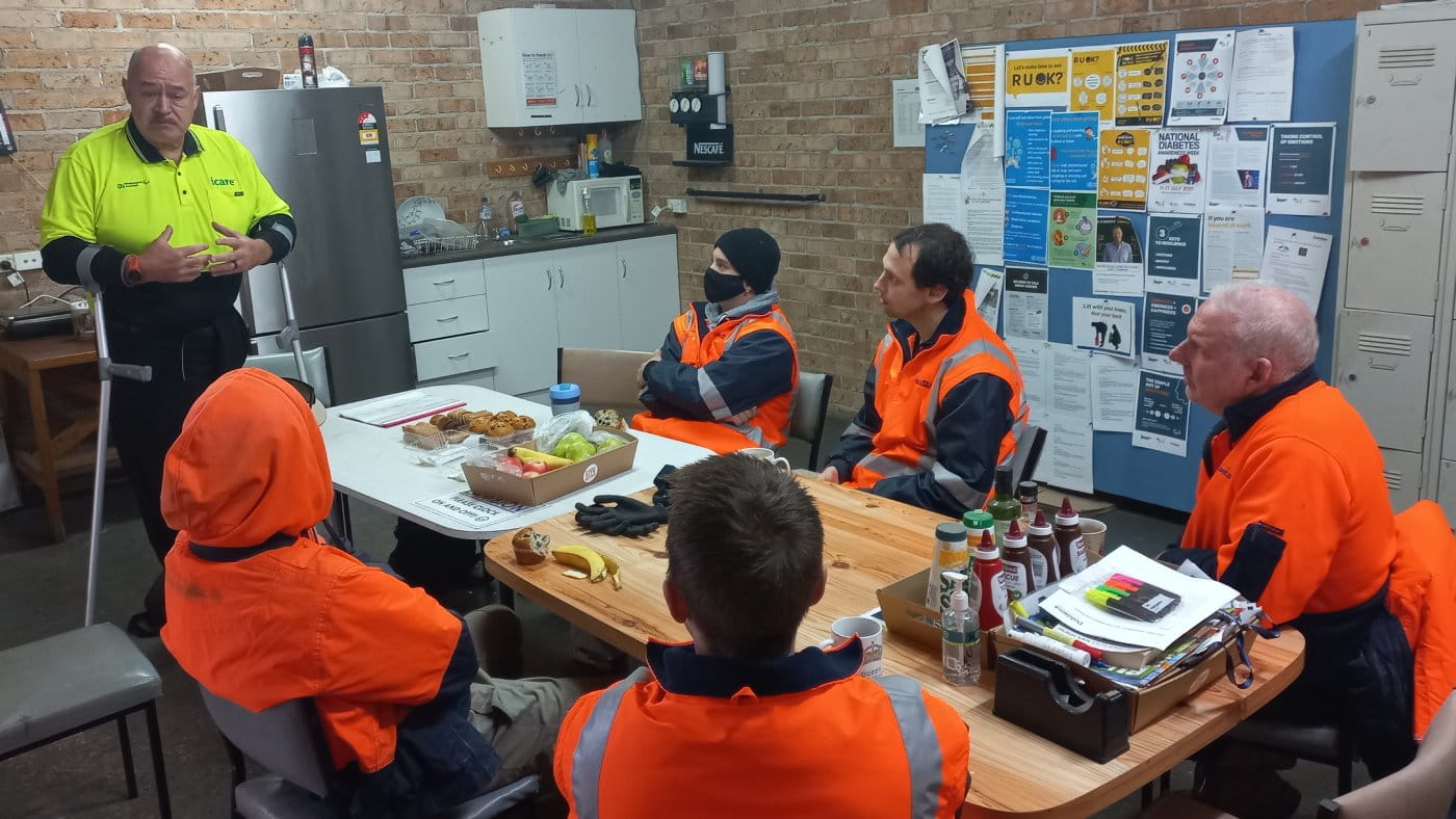 Kahi Puru wears a high-vis yellow shirt with icare and Paralympics Australia logo. He stands with the use of braces in a work breakout room with five workers in orange high-vis jumpers and jackets on, they sit at a table with a tray of biscuits and fruit in front of them. A small kitchen is visible and a pinup board is at the back with a number of items including RUOK, National Diabetes and safety messaging.