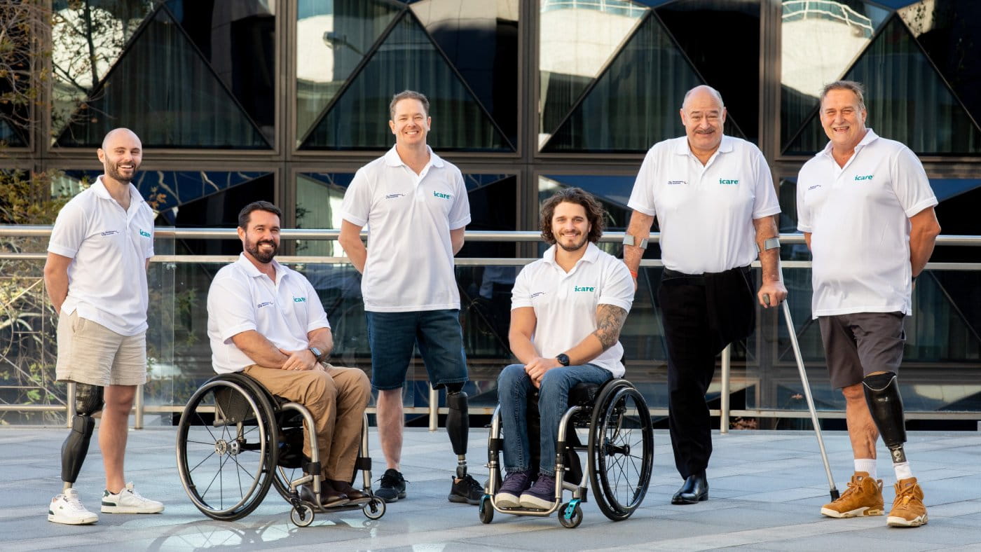 Six men are in the photo wearing white polo t-shirts with the icare logo. Two sit in wheelchairs, 3 have visible artificial legs and another stands with crutches.