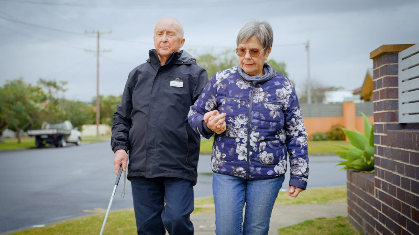 Doug Merrick walks on a suburban street with cane in right hand and wife Helen walks alongside holding his left hand.