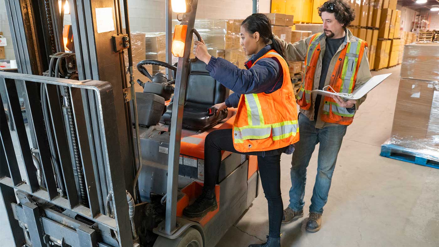 An industrial warehouse workplace safety topic. A safety supervisor or manager training a new employee on forklift safety. The trainer is holding a clipboard and explaining proper mounting of the forklift, with three points of contact.