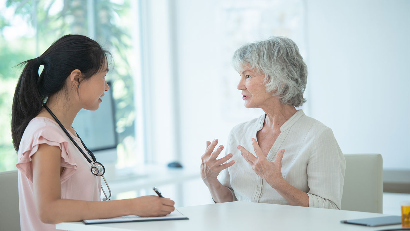 A female doctor of Asian descent listens to her elderly female Caucasian patient tell her about the problems that she has been having, both mentally and physically. The doctor takes notes so she can properly diagnose her.