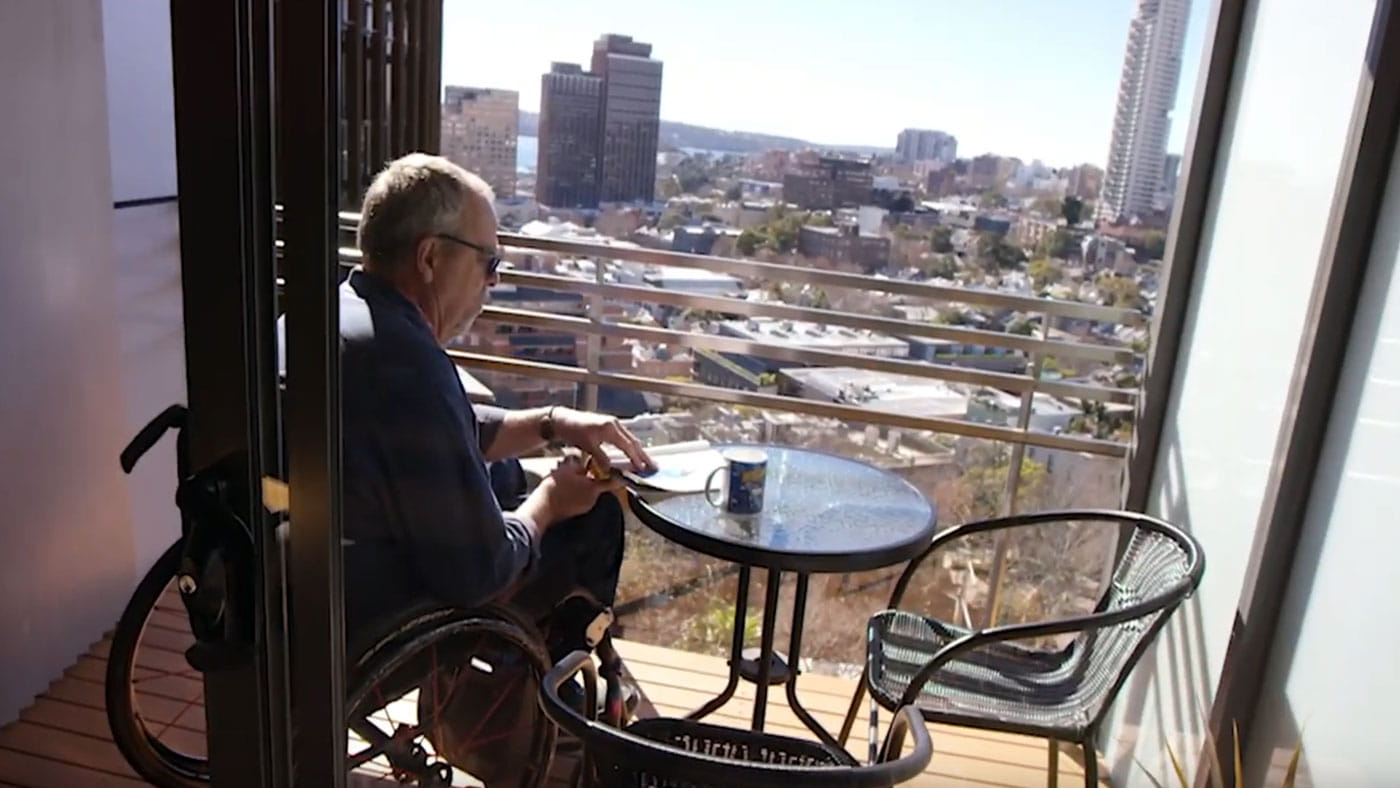 Man in wheelchair on balcony reading a magazine.